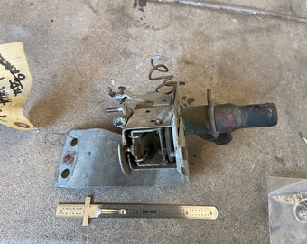 May include: A silver metal car part with a black rubber hose and a spring. The part has a rectangular base with two holes and a lever on the side. The part is on a gray concrete surface.