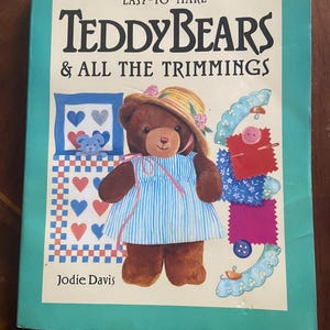 May include: A book titled "Easy-To-Make Teddy Bears & All The Trimmings" by Jodie Davis. The cover features a brown teddy bear wearing a blue and white striped dress and a straw hat. The bear is surrounded by fabric scraps and sewing supplies.