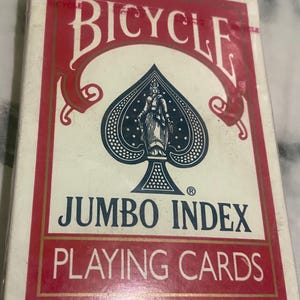 May include: A deck of Bicycle Jumbo Index playing cards in a white box with red borders. The front features the brand name in white, a large blue spade symbol containing a figure, and the text JUMBO INDEX PLAYING CARDS in blue and white.