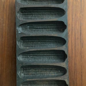 May include: A cast iron cornbread pan with seven corn cob-shaped molds. The pan is dark gray and sits on a wooden surface. The handle has a hole for hanging. A kitchenware item.