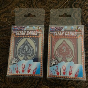 May include: Two decks of H2O Playing Cards in clear plastic cases. Each box features the text "H2O Playing Cards" and "Clear Cards" with a graphic of a spade and the letters J, Q, K, and A. One deck has a black spade, the other a red spade.