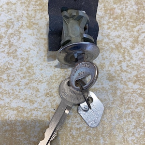 May include: A silver-colored car door lock assembly with two keys. The lock has a circular face and a black mounting plate. One key is a standard key, and the other is a small tag with text.