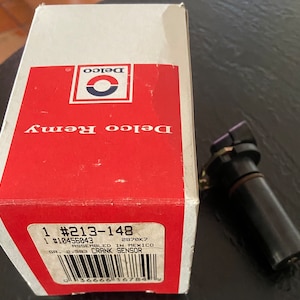 May include: A Delco Remy crank sensor and its packaging. The box is white and red with the Delco logo and text. The sensor is black with a cylindrical body. The box has a label with the part number and other details.