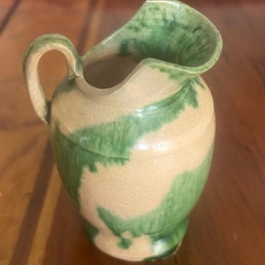 May include: A small, green and cream colored ceramic pitcher with a wide mouth and a handle. The pitcher has a rustic, handmade look.