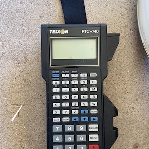 May include: A black Telxon PTC-740 handheld data terminal with a small display screen and a full alphanumeric keypad. The device has a black strap attached to the top and features various function keys and a red "ENTER" button.