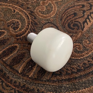May include: A white, rounded bottle stopper with a silver-colored threaded base. The stopper has a smooth, glossy finish. The background is a brown and black patterned fabric.