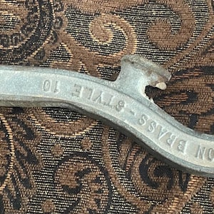 May include: A silver-colored Akron Brass Style 10 tool. The tool has a curved design with a hook on one end and a pointed end on the other. The text "AKRON BRASS - STYLE 10" is embossed on the tool.
