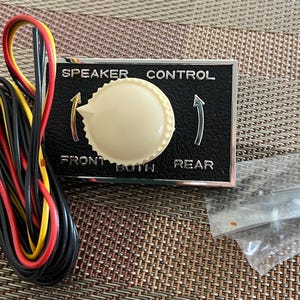 May include: A black speaker control knob with chrome trim. The knob has a white center and is labeled "SPEAKER CONTROL" with "FRONT", "BOTH", and "REAR" options.