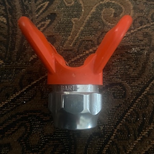 May include: A spray gun nozzle with a bright orange handle and a silver-colored metal base. The nozzle has a V-shaped handle and is designed for use with paint sprayers. The image shows the nozzle against a patterned background.