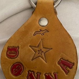 May include: A tan leather keychain with a silver key ring. The keychain has a star, a bird, and the word "DONNA" in red lettering.