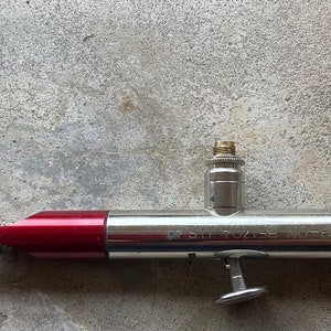 May include: A silver and red airbrush with a silver air tank. The airbrush has a red handle and a silver nozzle. The air tank has a silver cap and a black opening.