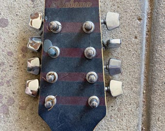 May include: A guitar headstock with a black and dark red striped finish featuring the word Alabama printed in gold script. The headstock includes eight chrome tuning pegs arranged in two columns of four, with metal posts and some remaining string ends.