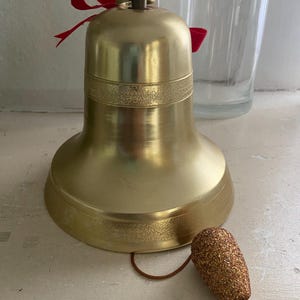 May include: A gold-colored bell with a red ribbon tied to the top. The bell has a decorative band around the middle and a small, textured clapper. The bell is likely used for decoration or as a holiday ornament.