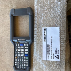 May include: A black handheld barcode scanner with a keypad and a screen. The scanner has the Intermec logo on it. The scanner is sitting on a gray surface. The text on the label reads: ITEM: 714-766-004FRE DESC: FRE, COVER, TOP ALPHA-NUMERIC ASSY BOMREV: B STATIC SENSITIVE DEVICE Honeywell