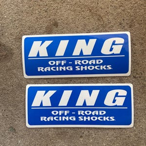 May include: Two rectangular blue stickers with white borders. The text KING is printed in large white capital letters above the text OFF-ROAD RACING SHOCKS in smaller white capital letters. The stickers have a glossy finish and rounded corners.