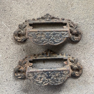 May include: Two dark brown, weathered metal drawer pulls featuring an ornate, raised floral and scrollwork pattern. Each pull has a rectangular central slot for a label and two circular mounting holes on the sides with a textured, aged finish.