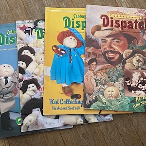May include: A stack of four Cabbage Patch Kid magazines. The top magazine is titled "Collector's Dispatch" and features a portrait of a man with a beard. The second magazine is titled "Cabbage Patch Kids Dispatch" and features a Cabbage Patch Kid doll holding a paint palette. The third magazine is titled "Cabbage Patch Kids Dispatch" and features a group of Cabbage Patch Kid dolls. The bottom magazine is titled "Cabbage Patch Kids Dispatch" and features a Cabbage Patch Kid doll wearing a trench coat.