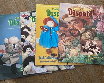 May include: A stack of four Cabbage Patch Kid magazines. The top magazine is titled "Collector's Dispatch" and features a portrait of a man with a beard. The second magazine is titled "Cabbage Patch Kids Dispatch" and features a Cabbage Patch Kid doll holding a paint palette. The third magazine is titled "Cabbage Patch Kids Dispatch" and features a group of Cabbage Patch Kid dolls. The bottom magazine is titled "Cabbage Patch Kids Dispatch" and features a Cabbage Patch Kid doll wearing a trench coat.