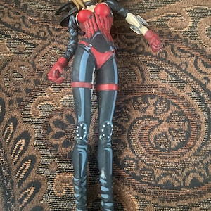 May include: A black and red action figure of a female character with blonde hair, wearing a black leather jacket, red and black pants, and black boots. The figure has a robotic arm and is holding a red weapon.
