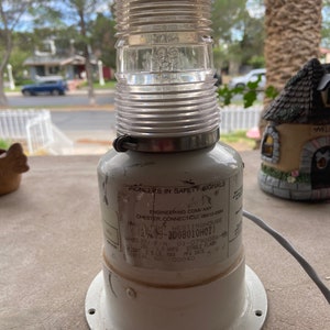 May include: A vintage safety signal light with a clear, ribbed, cylindrical top and a white base. The light has a metal band and a label with text including "Pioneers in Safety Signals" and "Whelen".