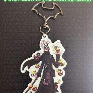 May include: A 3-inch glow-in-the-dark keychain featuring a character in a long dark coat surrounded by small ducks. The text reads: SCREAM FORTRESS MERASMUS & MERCENARY DUCKS 3 INCH GLOW IN THE DARK KEYCHAIN. It has a bat-shaped metal clasp at the top.