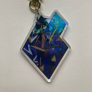 Puede incluir: Un llavero iridiscente azul y amarillo con un personaje que sostiene una espada y una pistola. El personaje está rodeado de un patrón geométrico azul y amarillo. El llavero está unido a un cierre dorado en forma de estrella.