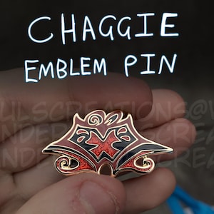 May include: A gold-colored pin with a red and black emblem. The emblem has a stylized design with a red center and black outlines. The pin is a Chaggie Emblem Pin.