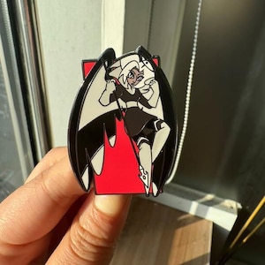 May include: Black and white enamel pin featuring a cartoon character with wings, a black dress, and a red background.