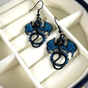 Pode incluir: Um par de brincos de drag&atilde;o pretos com detalhes em glitter azul. Os brincos apresentam um design detalhado de drag&atilde;o com asas abertas e cauda enrolada. S&atilde;o apresentados numa caixa de joias branca.