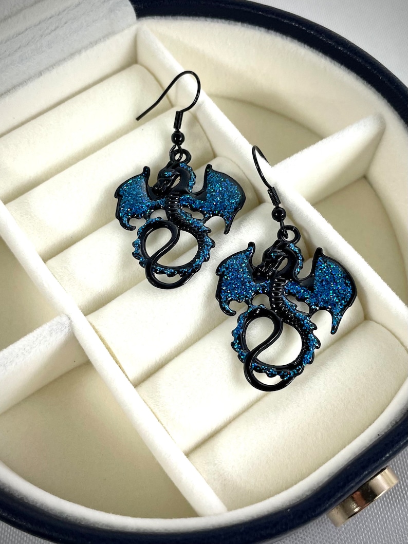 Pode incluir: Um par de brincos de drag&atilde;o pretos com detalhes em glitter azul. Os brincos apresentam um design detalhado de drag&atilde;o com asas abertas e cauda enrolada. S&atilde;o apresentados numa caixa de joias branca.
