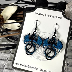 Pode incluir: Brincos de drag&atilde;o pretos com asas e detalhes em glitter azul. Os brincos s&atilde;o exibidos em um cart&atilde;o branco com o texto "Daring Impressions" e um endere&ccedil;o de site. Ganchos de brinco pretos.