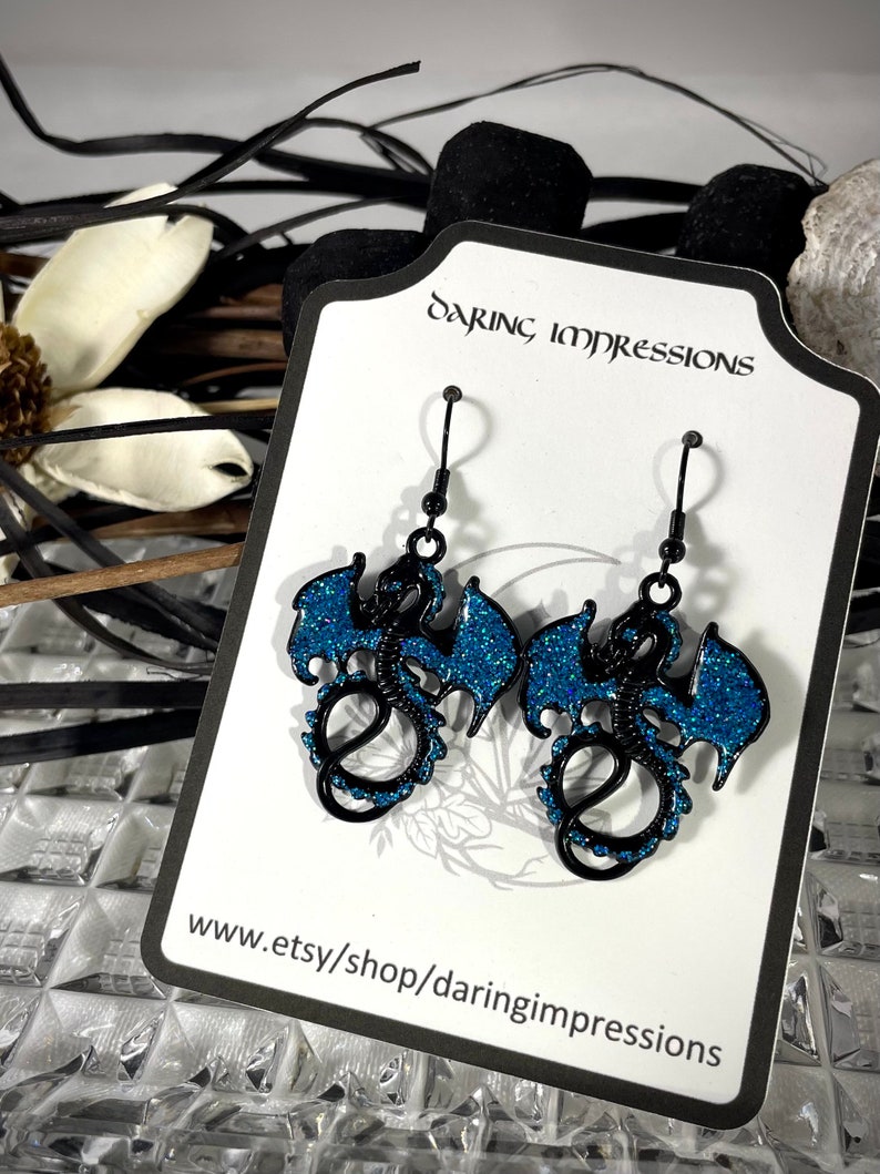 Pode incluir: Brincos de drag&atilde;o pretos com asas e detalhes em glitter azul. Os brincos s&atilde;o exibidos em um cart&atilde;o branco com o texto "Daring Impressions" e um endere&ccedil;o de site. Ganchos de brinco pretos.