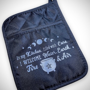 May include: Black quilted oven mitt with a loop for hanging. The mitt features the text "In my Kitchen, filled with Care. I WELCOME Water, Earth, Fire & Air" with a cauldron and moon phases design.