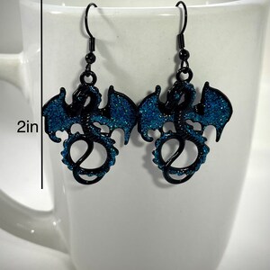 Pode incluir: Brincos de drag&atilde;o pretos com asas e detalhes em glitter azul. Cada brinco tem aproximadamente 5 cm de comprimento, apresentando um design detalhado de drag&atilde;o com fecho de gancho. Os brincos s&atilde;o exibidos em um fundo branco.