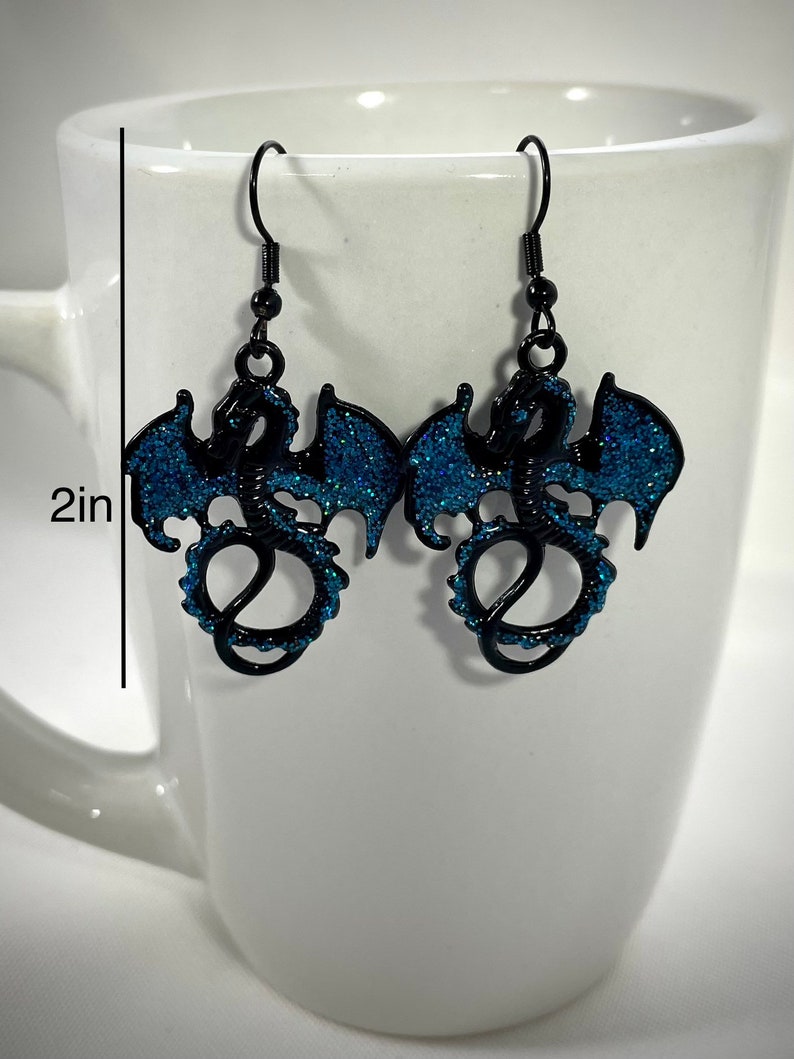 Pode incluir: Brincos de drag&atilde;o pretos com asas e detalhes em glitter azul. Cada brinco tem aproximadamente 5 cm de comprimento, apresentando um design detalhado de drag&atilde;o com fecho de gancho. Os brincos s&atilde;o exibidos em um fundo branco.