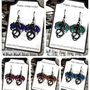 Pode incluir: Cinco pares de brincos em forma de drag&atilde;o em v&aacute;rias cores: roxo, verde-azulado, vermelho, laranja e azul. Cada par apresenta um contorno preto com asas e corpo preenchidos com glitter. Os brincos s&atilde;o exibidos em cart&otilde;es brancos com texto.