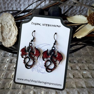 May include: Black dragon earrings with red glitter wings and accents. The earrings are displayed on a white card with the text "Daring Impressions" and the Etsy shop URL.