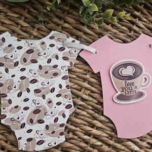 May include: Baby shower banner featuring onesie cutouts. One onesie is pink with a coffee cup graphic and the text "Love You a Latte." Other onesies have a coffee bean and cup pattern in brown and white. Ribbons are attached for hanging.