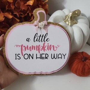 Może przedstawiać: Tablica w kształcie dyni z napisem "a little pumpkin is on her way". Tablica ma r&oacute;żowo-złotą brokatową obw&oacute;dkę i małą kokardkę. Biała dynia ze złotą łodygą i pomarańczowa dynia dekoracyjna znajdują się w tle.