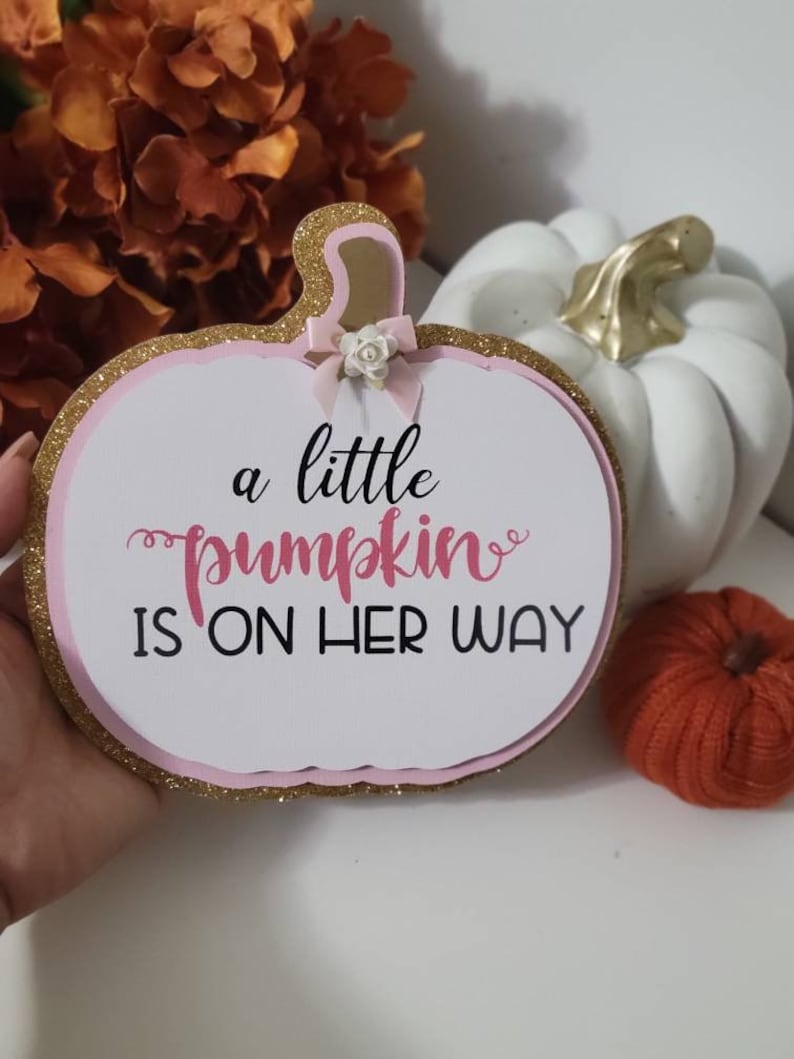 Może przedstawiać: Tablica w kształcie dyni z napisem "a little pumpkin is on her way". Tablica ma r&oacute;żowo-złotą brokatową obw&oacute;dkę i małą kokardkę. Biała dynia ze złotą łodygą i pomarańczowa dynia dekoracyjna znajdują się w tle.