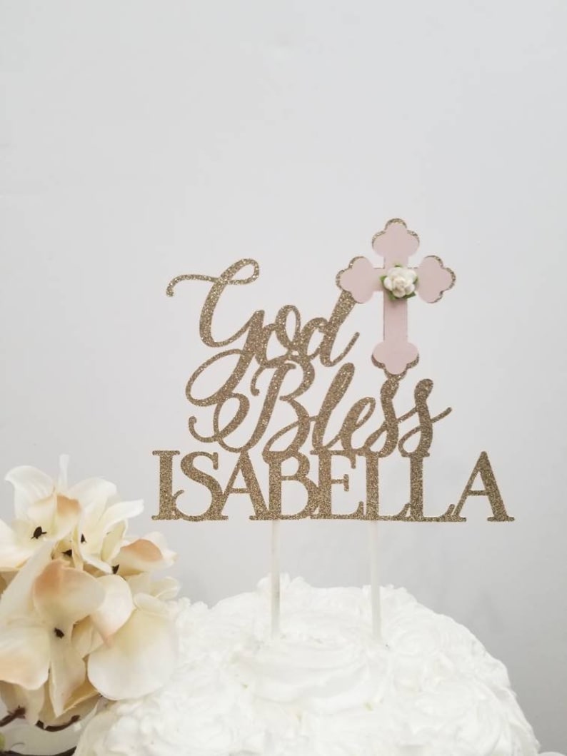 May include: Gold glitter cake topper with the text "God Bless Isabella" and a pink cross with a white flower.