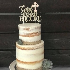 May include: Two-tiered, semi-naked cake decorated with hessian ribbon and succulents. The cake is topped with a glitter cake topper that says "God Bless Brooke" and features a pink cross.