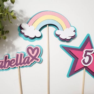 May include: A set of colorful birthday cake toppers. Includes a rainbow, a star with the number 5, and a name topper that says "Isabella". Each topper is multi-layered with a variety of colors.