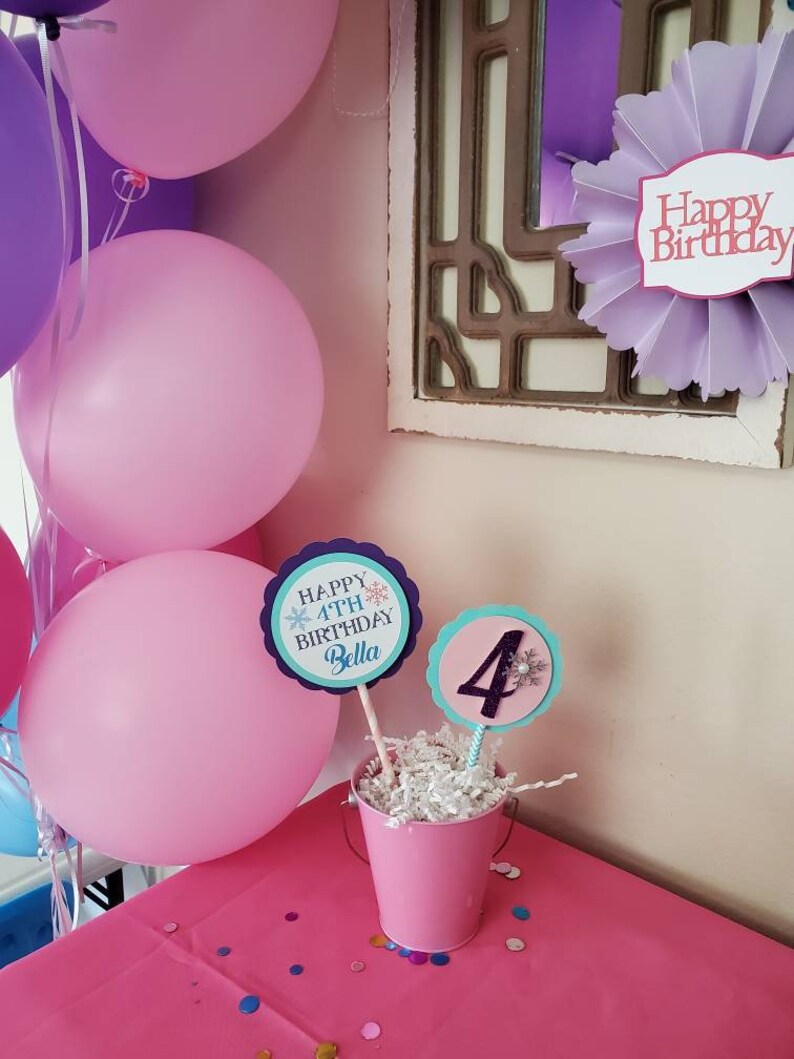 May include: A pink birthday party scene with balloons, decorations, and a table. The table has a pink tablecloth and a small pink bucket with birthday signs that read "Happy 4th Birthday Bella" and the number "4".