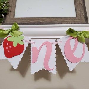 May include: A white banner with pink and red cutouts that spell out "one". The banner has green bows and a red strawberry cutout.