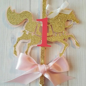 May include: Gold glitter carousel horse cake topper with a pink bow and a red number one. The topper is on a striped stick.