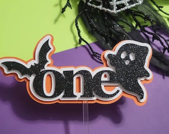 May include: Orange and white cake topper with the word "one" in black glitter, a black bat, and a black ghost.