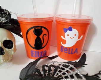 May include: Two orange plastic cups with lids and straws. One cup has a black cat design and the name "Eliza" in purple. The other cup has a white ghost design and the name "Bella" in purple.