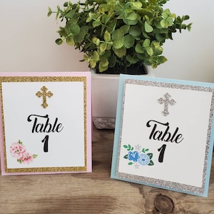 May include: Two table number cards with a gold glitter border and a silver glitter border. The cards are white with a black cross, black text that says "Table 1", and floral designs. The card with the gold border has pink flowers and the card with the silver border has blue flowers.