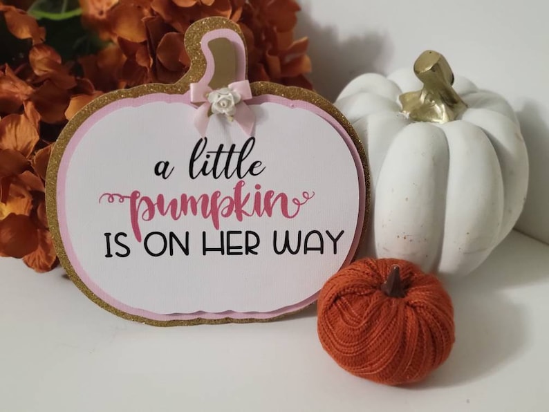 Może przedstawiać: Ozdobny znak w kształcie dyni z napisem "a little pumpkin is on her way". Znak ma złotą, błyszczącą obw&oacute;dkę i r&oacute;żowe akcenty. Na zdjęciu znajduje się r&oacute;wnież mała biała dynia i pomarańczowa dynia z tkaniny.
